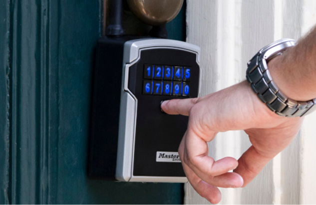 A finger pressing the illuminated keypad of a wall-mounted Master Lock lock box